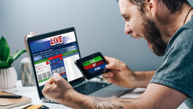 Young attractive bearded man showing sincere excitement about his favorite team victory. Guy being happy winning a bet in online sport gambling application on his mobile phone.