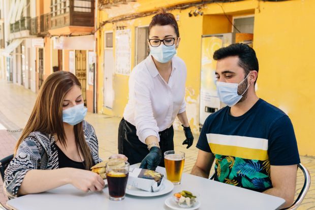 two people wearing masks being served by a waiter wearing a mask at an outdoor restaurant