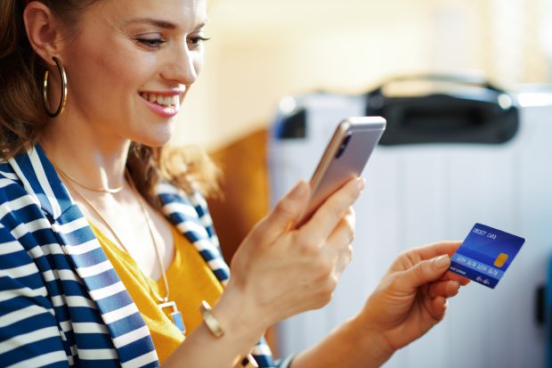 smiling young tourist woman in striped jacket with credit card buying airplane tickets online on a smartphone while sitting near couch and trolley bags at modern home in sunny hot summer day.